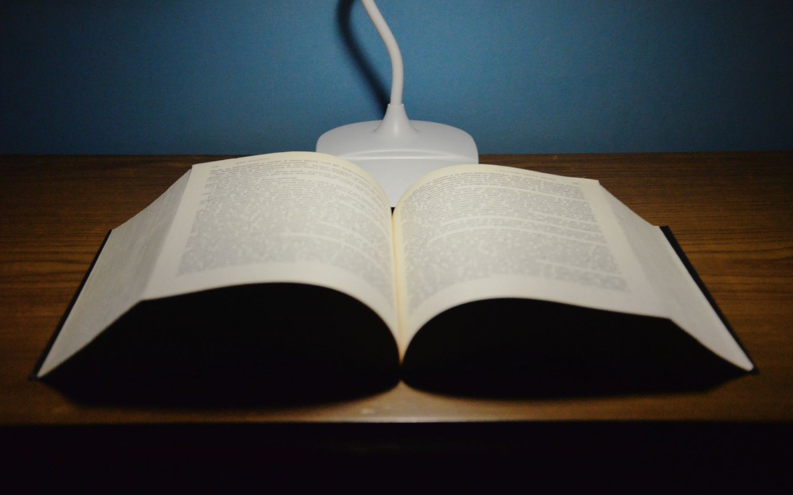 An open thick book on a wooden desk beneath a small white desk lamp