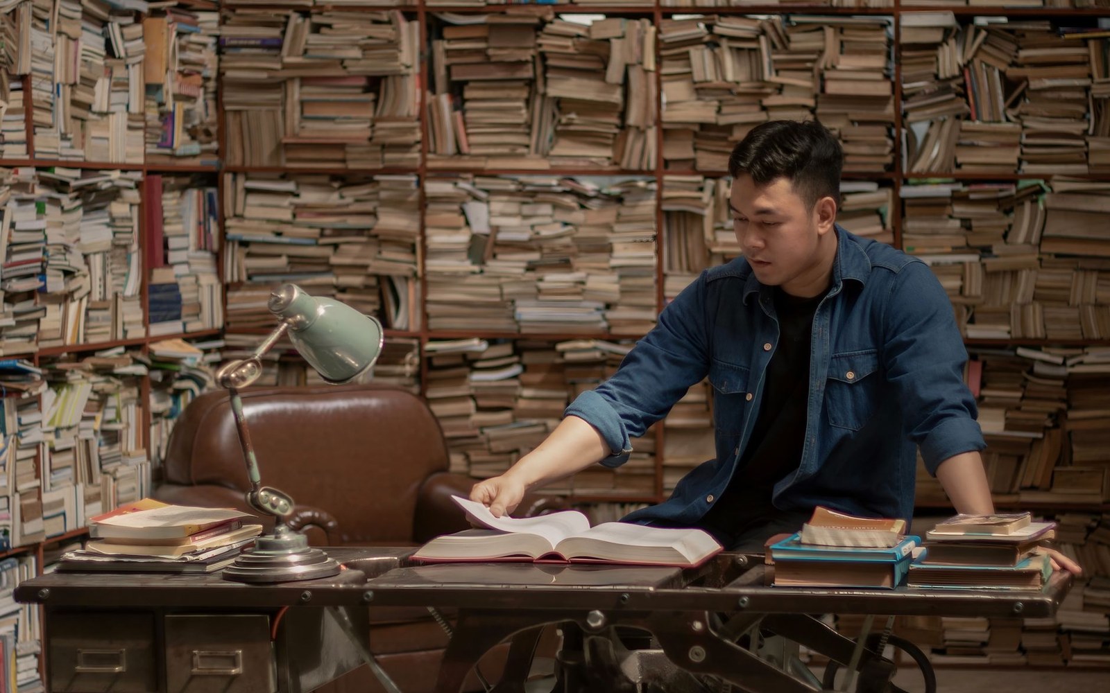 Man leaning on a desk flipping through a book in a room stacked with books