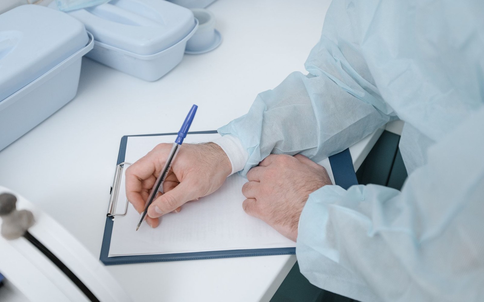 A gloved clinician writes notes on a clipboard beside sterile trays on a table