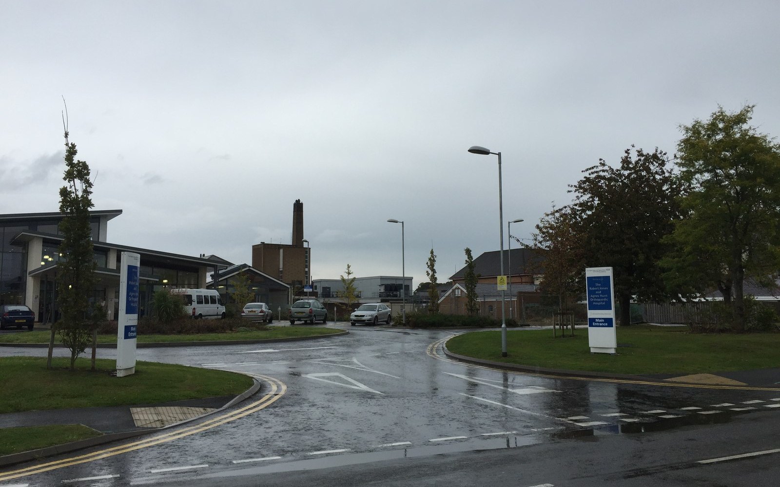 Wet entrance road leading to hospital buildings with signage and a tall brick chimney
