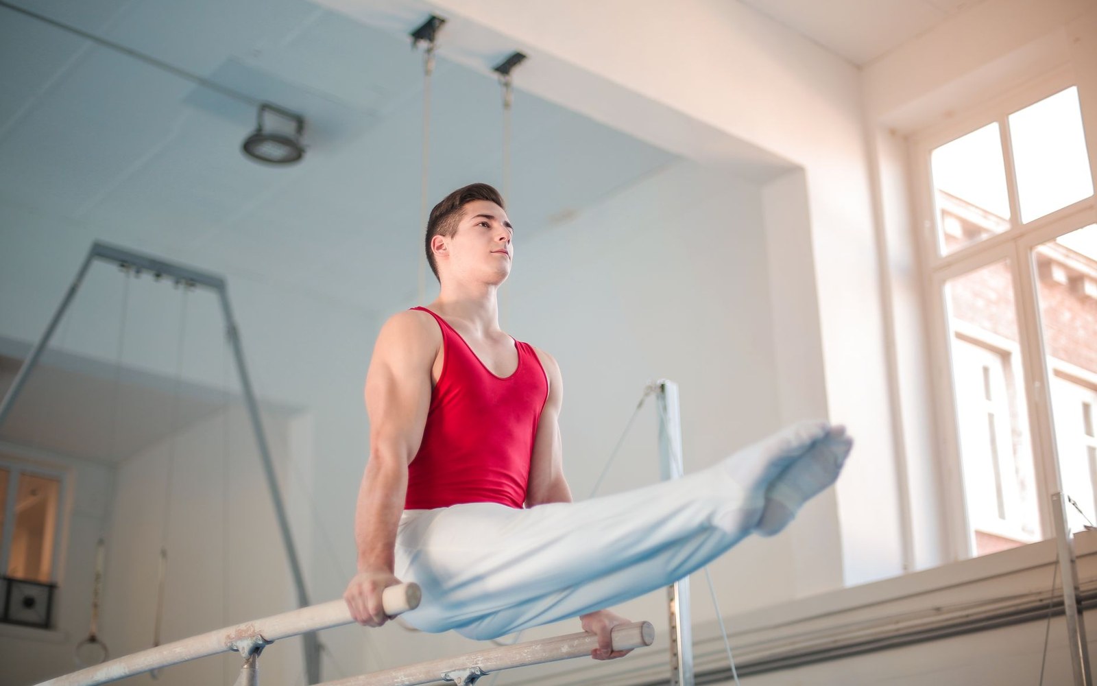 A male gymnast in a red leotard balances on parallel bars with legs extended