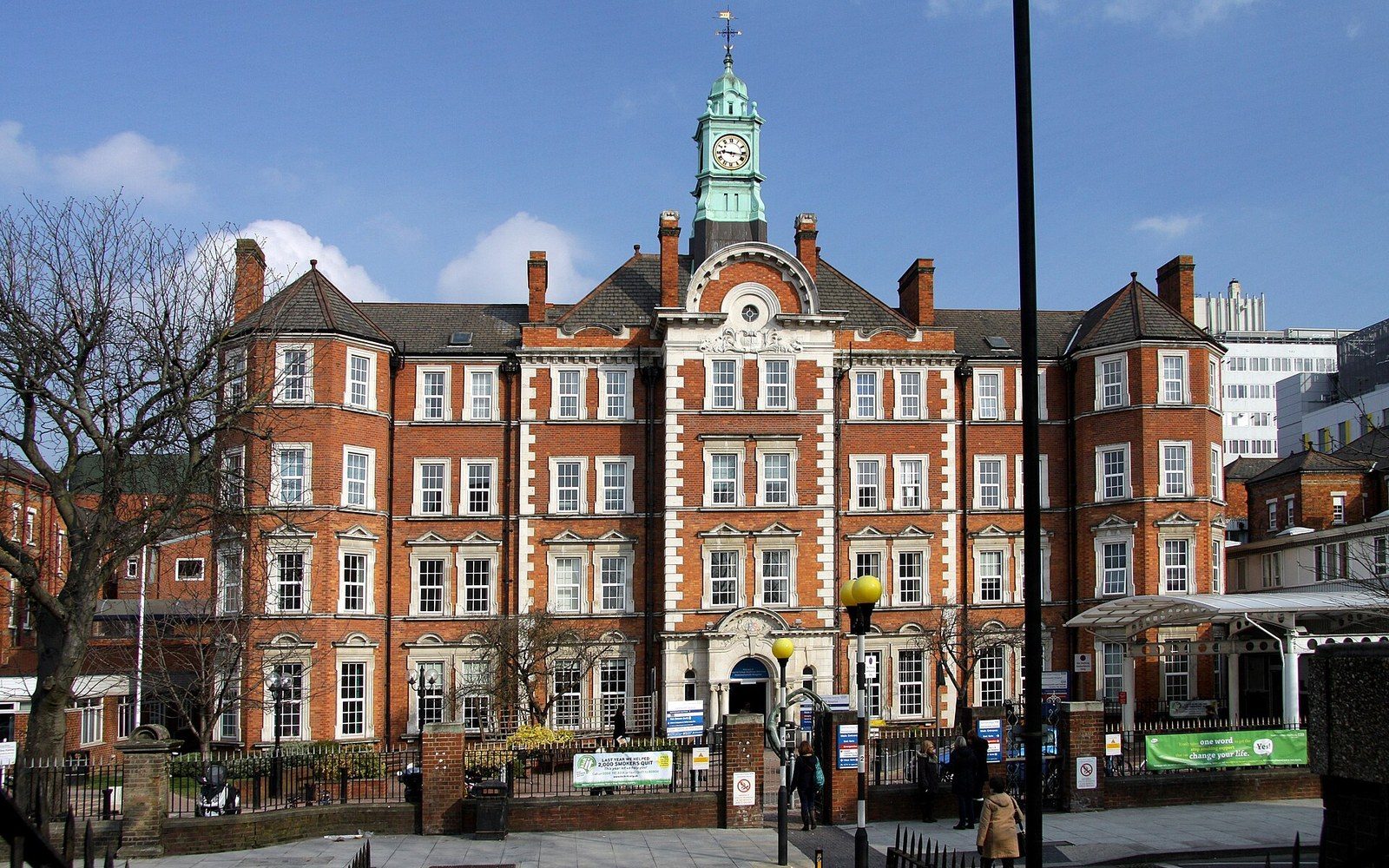 Redbrick hospital building with a clock tower and green cupola under a blue sky