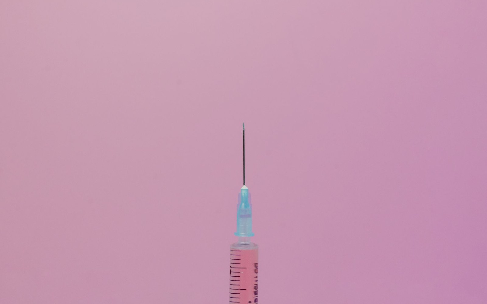 A syringe with a needle stands upright against a pink background