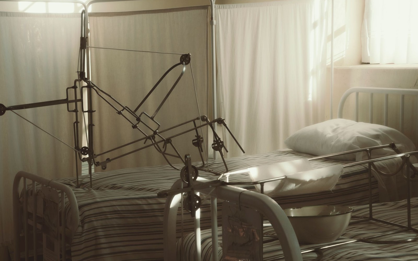 Old hospital bed with a metal traction frame beside curtained windows in soft light