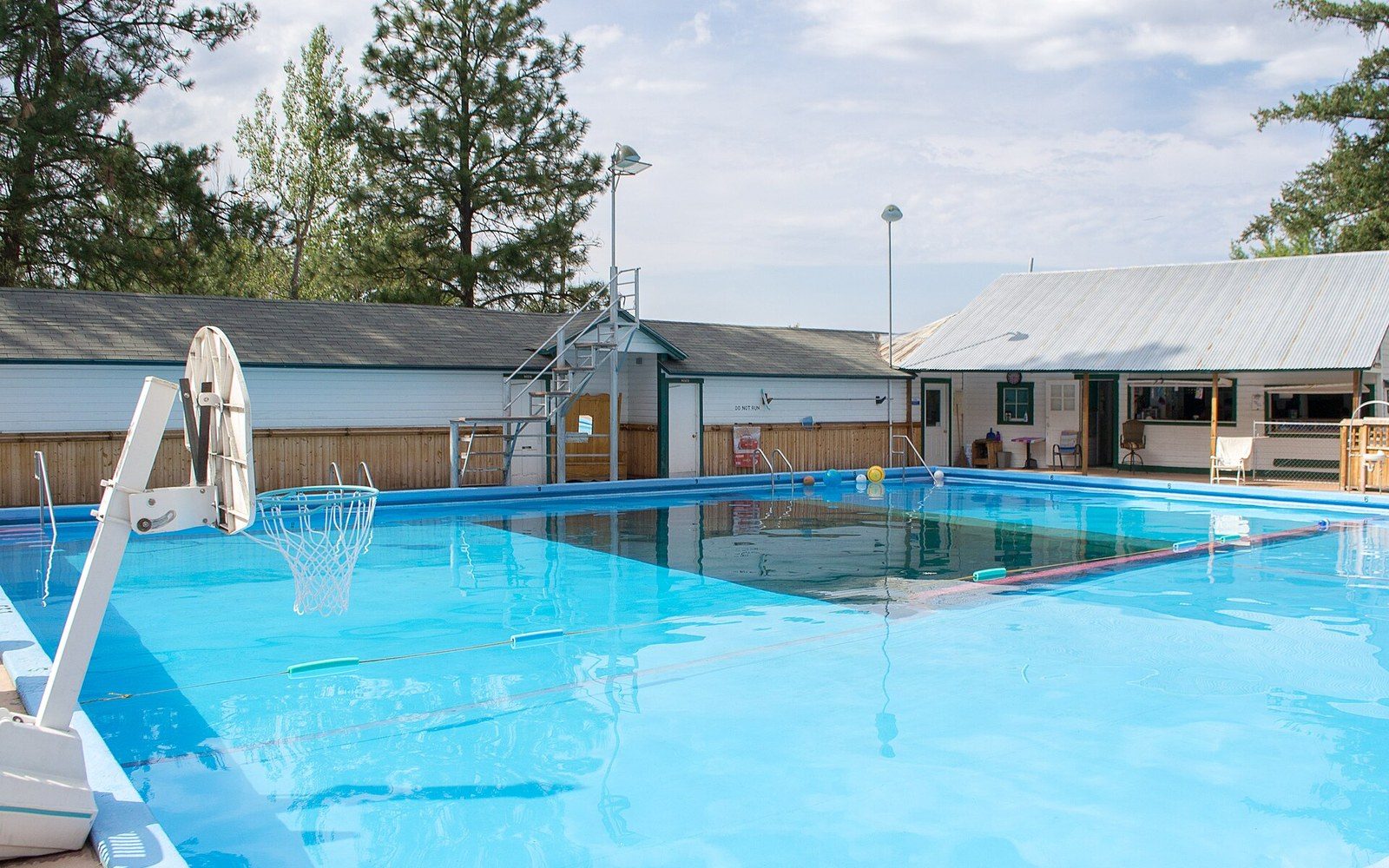 Outdoor swimming pool with a basketball hoop, lifeguard chair and poolside cottages under a partly cloudy sky