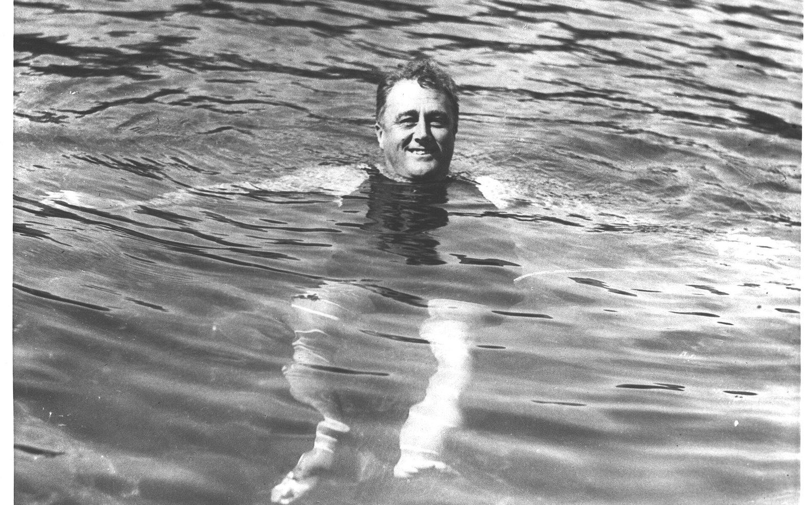 Franklin Roosevelt in the pool at Warm Springs, Georgia, adult figures in the water around him, the colonnade visible behind — landscape, full-bleed homepage lead